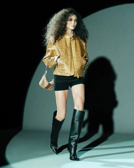 Fashion model in golden jacket, mini shorts, and knee-high boots, posing with a Gucci handbag in a studio spotlight.