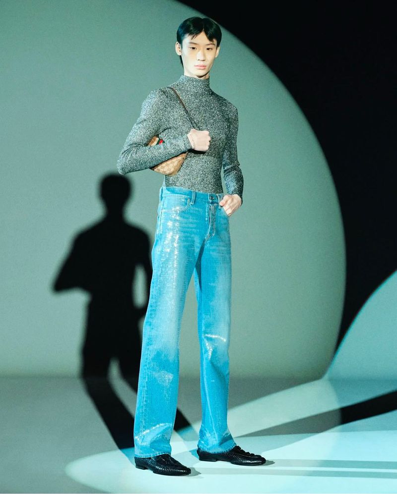 Full shot of a slender adult male in a turtleneck and shiny pants, holding a small purse with a shadow on a blue background.