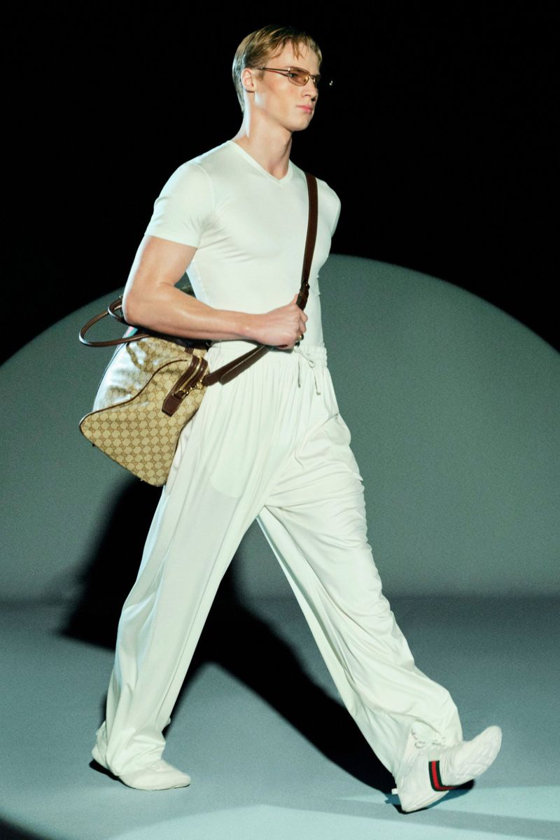 Fashion model walks runway in white outfit, shoulder bag, and sneakers during a show, highlighting modern design and style.
