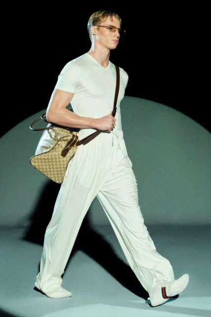 Fashion model walks runway in white outfit, shoulder bag, and sneakers during a show, highlighting modern design and style.
