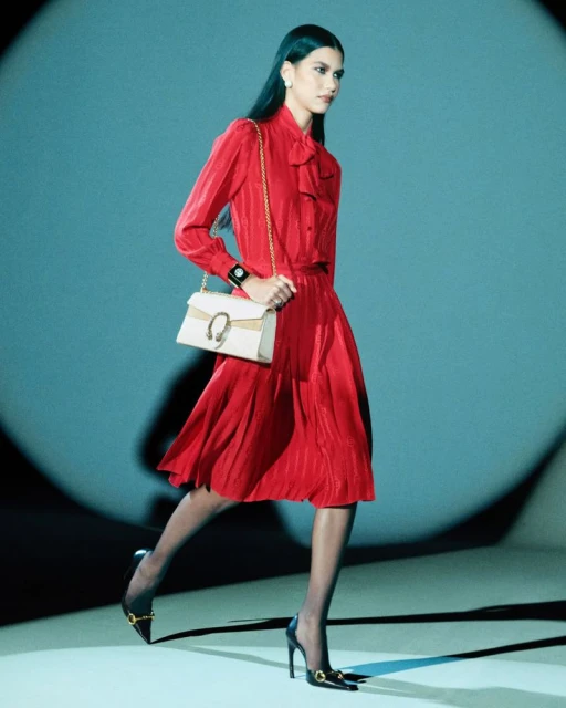 Fashion model in a red dress and black heels walks in a studio with a designer handbag, long dark hair, and light blue background.