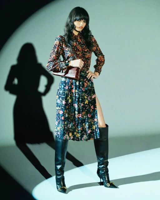 A woman poses indoors, wearing a floral long-sleeved blouse and skirt with a thigh-high slit, paired with black leather heeled boots.