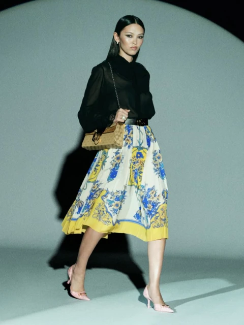 An Asian woman in a sheer black top and floral skirt poses with a handbag in a studio with a light shining on her. Fashion shot.
