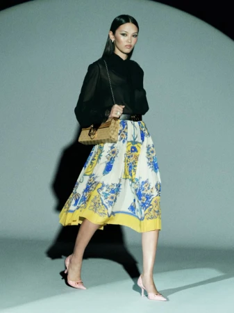 An Asian woman in a sheer black top and floral skirt poses with a handbag in a studio with a light shining on her. Fashion shot.