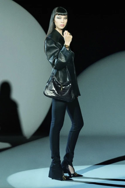 Runway fashion shot featuring an adult female model in black jacket, fitted pants, and high heels, accessorized with a black purse.
