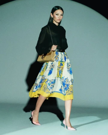 Model walking in a black blouse, yellow patterned midi skirt, pink kitten heels, and carrying a tan shoulder bag in a spotlight.