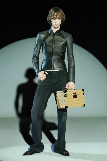 A fashion model poses in a dark leather jacket and black pants, holding a patterned briefcase in a studio setting, creating a high-fashion editorial image.