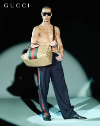 Shirtless male model wearing dark sunglasses and holding a large monogram duffel bag under a spotlight.