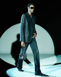 Black male model in a tailored dark blazer, trousers, and sunglasses walking with a small handbag in a circular studio spotlight.