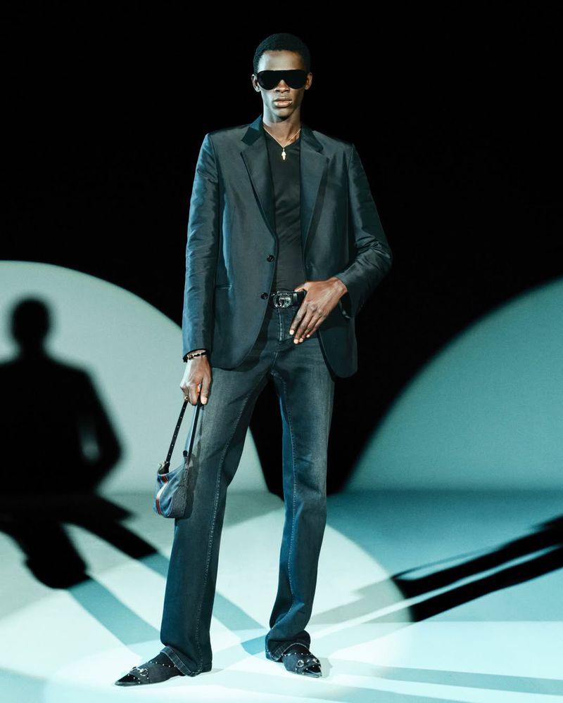 Black man wearing dark sunglasses, dark blazer, and denim jeans, holding a small handbag against a bright stage backdrop.