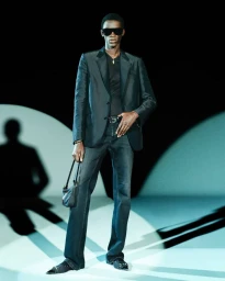 Black man wearing dark sunglasses, dark blazer, and denim jeans, holding a small handbag against a bright stage backdrop.