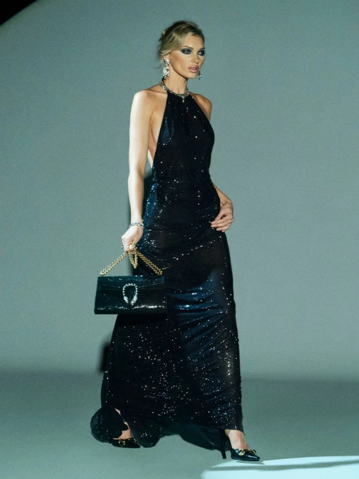 Model Elsa Hosk walks wearing a black sequined halter gown and holding a black handbag with a chain strap against a grey studio backdrop.