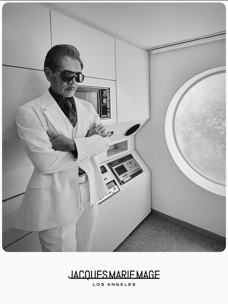 Monochrome portrait of an older Asian man in a white suit, wearing sunglasses indoors with a futuristic control panel and circular window.