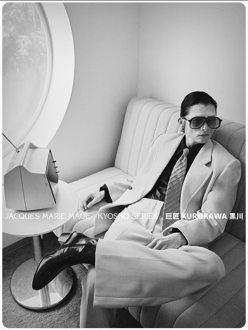 Black and white studio shot of a man in a light-colored suit and dark sunglasses, seated on a striped couch near an old television.