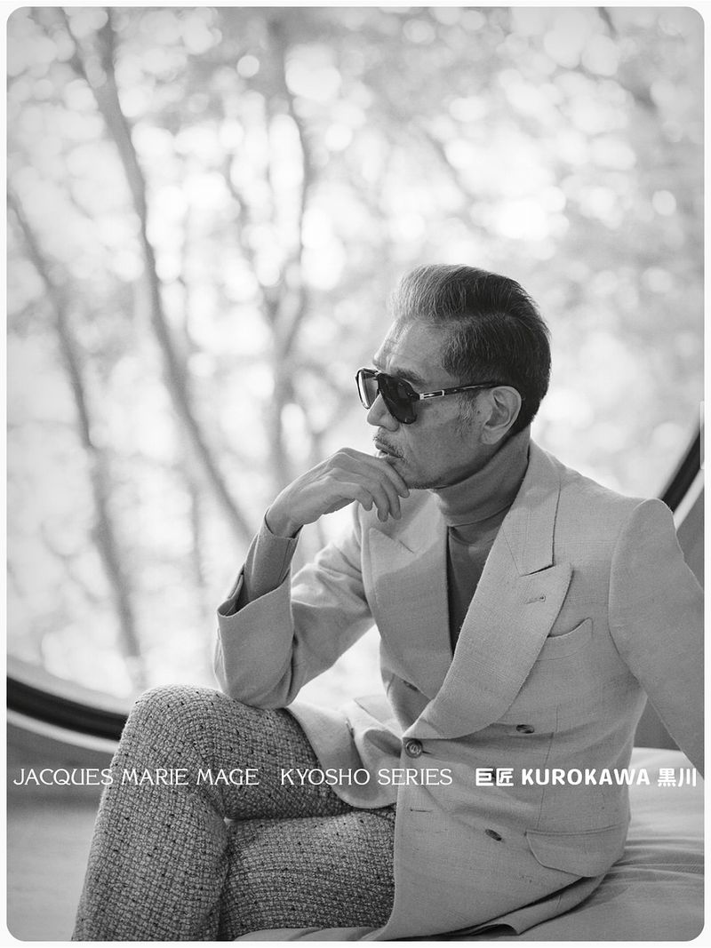 Mature East Asian man wearing Jacques Marie Mage sunglasses and a tailored double-breasted suit jacket by a round window.