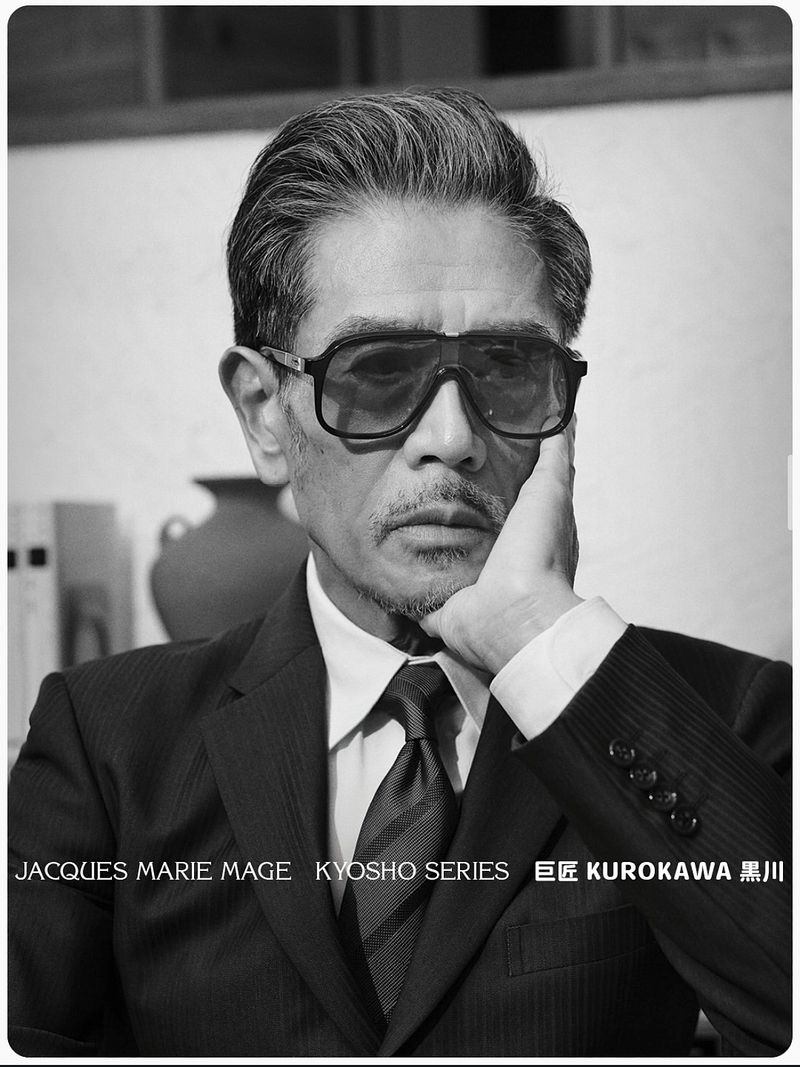 East Asian man in a dark suit, tie, and bold Jacques Marie Mage sunglasses resting his chin on his hand in black and white.
