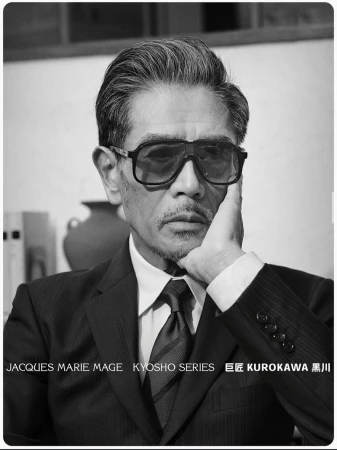 East Asian man in a dark suit, tie, and bold Jacques Marie Mage sunglasses resting his chin on his hand in black and white.