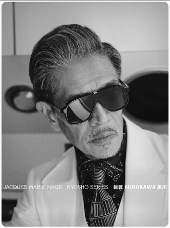 Mature East Asian man in oversized black sunglasses, white suit jacket, patterned cravat, and tie in black and white.