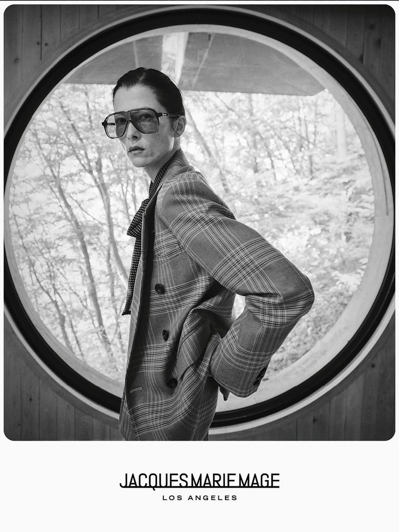 Monochrome portrait of a stylish adult with sunglasses and plaid blazer, posing near a round window with a forest view.