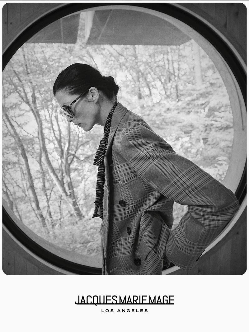 Black and white portrait of a stylish adult woman wearing sunglasses and a plaid blazer posing inside a building in front of a window.