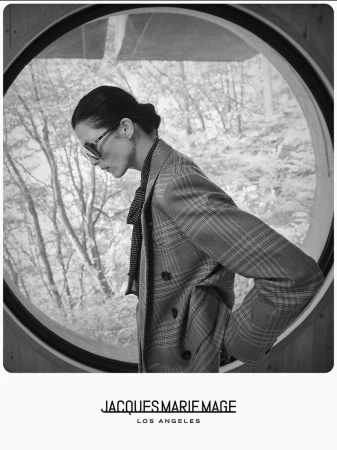 Black and white portrait of a stylish adult woman wearing sunglasses and a plaid blazer posing inside a building in front of a window.