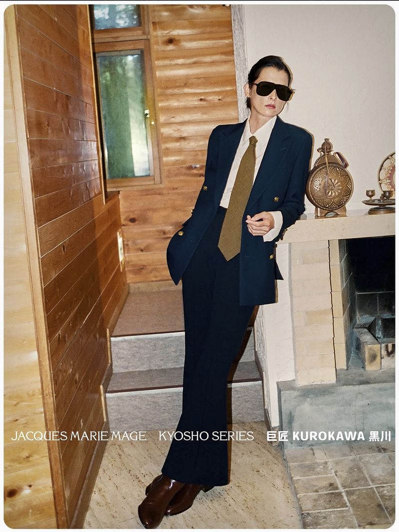 A woman in a dark suit, sunglasses, and tie poses in an indoor setting near a fireplace and wooden wall, creating a fashion-forward portrait.