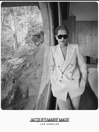 Man wearing dark sunglasses and a double-breasted suit jacket standing by a curved window in a wood-paneled room.