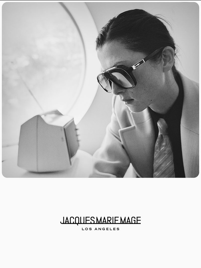 Black and white image of a person wearing aviator sunglasses and a blazer, looking down next to a retro television set.
