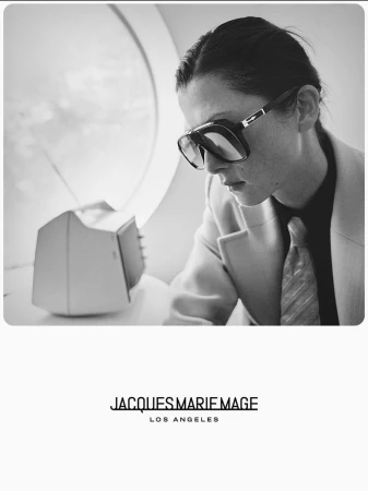 Black and white image of a person wearing aviator sunglasses and a blazer, looking down next to a retro television set.