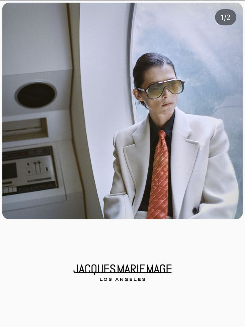Adult woman poses wearing sunglasses, a cream-colored suit, a black shirt, and a patterned orange tie against a modern background.