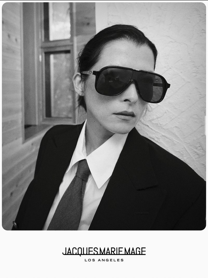 Model in oversized dark sunglasses, black suit jacket, white collared shirt, and necktie posing beside a window.