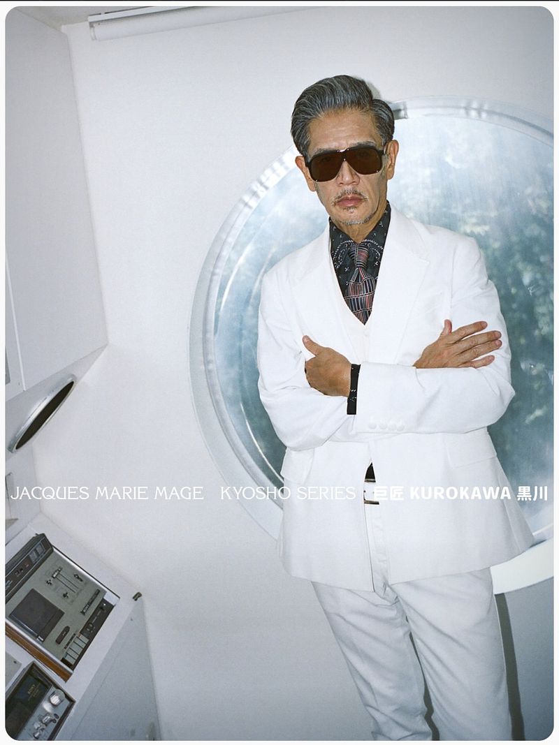 Stylish older Asian man in a white suit and sunglasses poses in a modern, well-lit interior with retro electronic equipment.