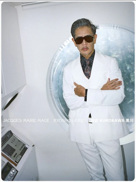 Stylish older Asian man in a white suit and sunglasses poses in a modern, well-lit interior with retro electronic equipment.
