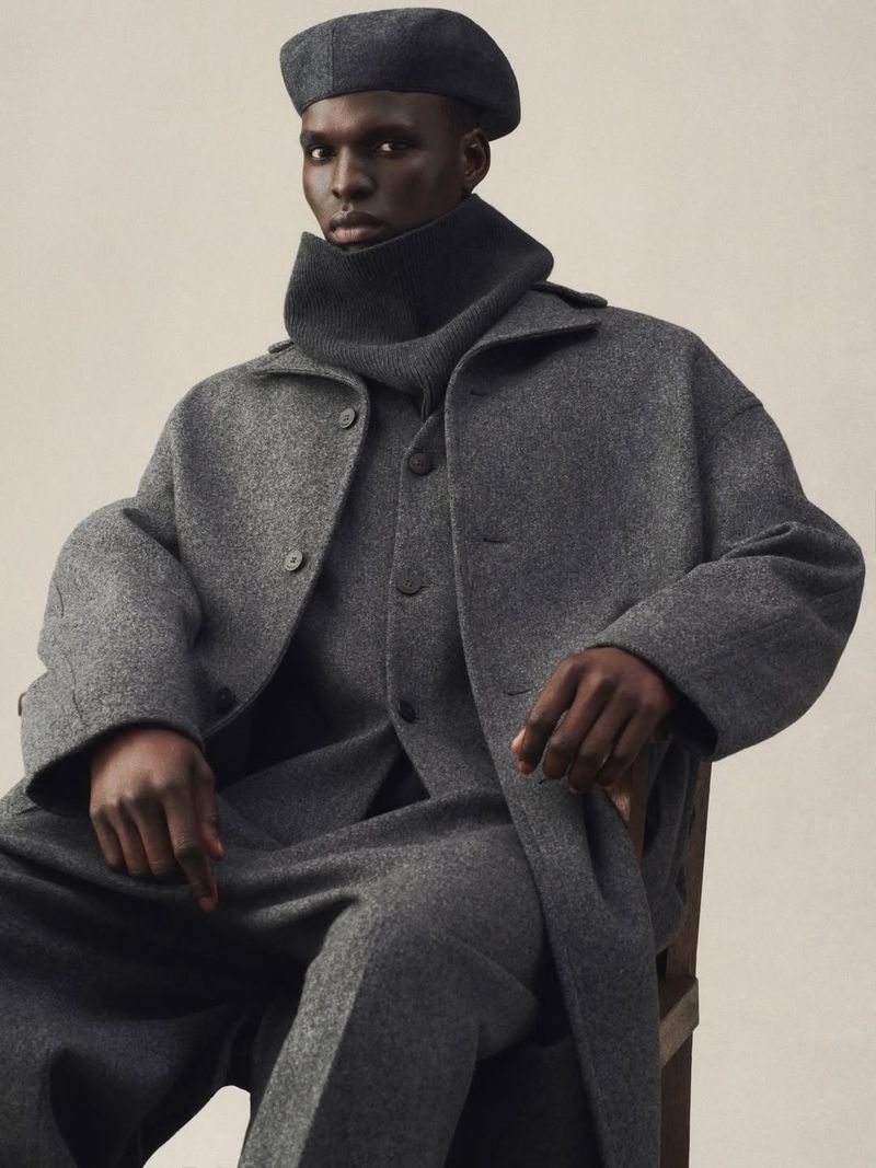 An adult male poses indoors while wearing a gray wool coat, sweater, beret, and trousers, sitting on a chair against an off-white backdrop.