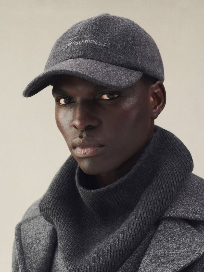 Close-up shot of a Black male model wearing a grey wool cap and matching wool overcoat against a neutral backdrop. Fashion editorial.