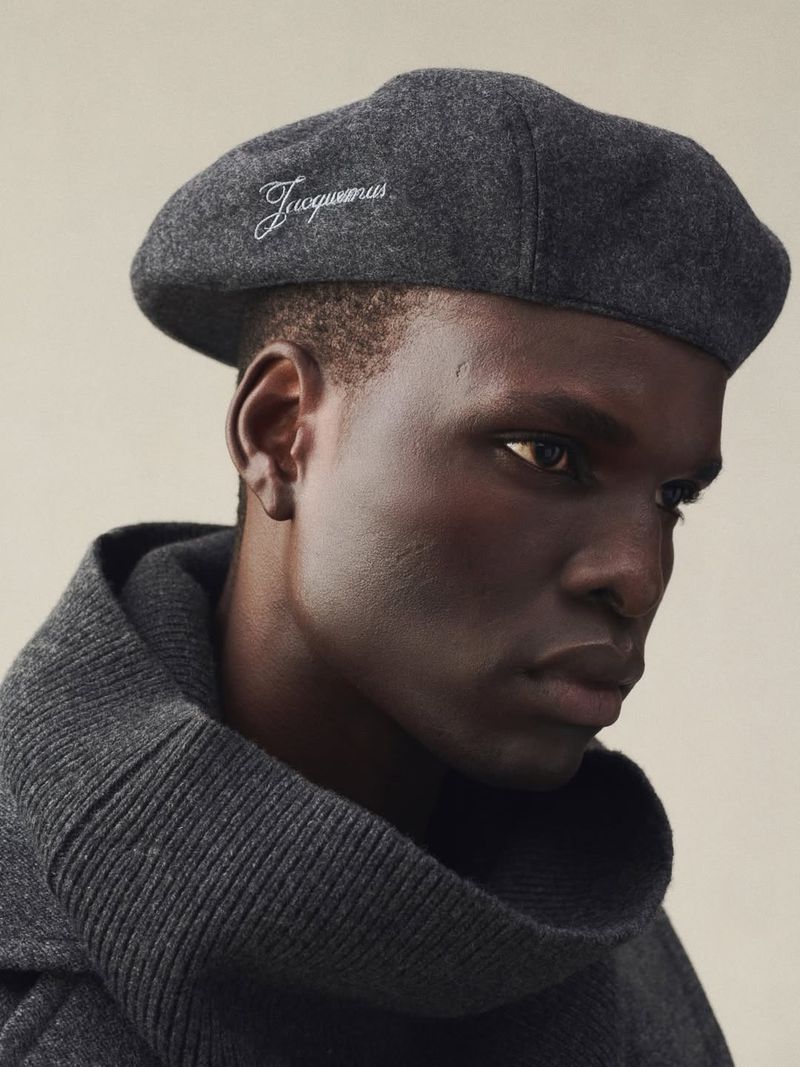 Portrait of a dark-skinned male model wearing a grey beret with brand text and grey high-necked sweater against a neutral backdrop.