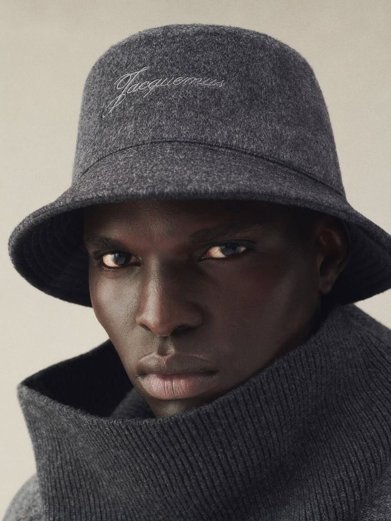 Close-up of a dark-skinned man wearing a grey bucket hat with the Jacquemus logo and a matching turtleneck sweater, neutral background.