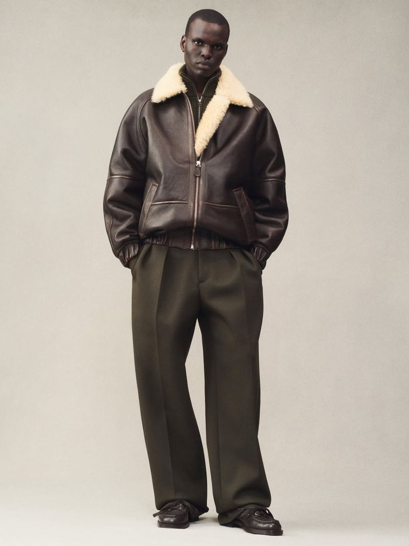 Full-length studio portrait of an adult Black male model wearing a dark brown leather aviator jacket with khaki-green trousers and dark shoes.