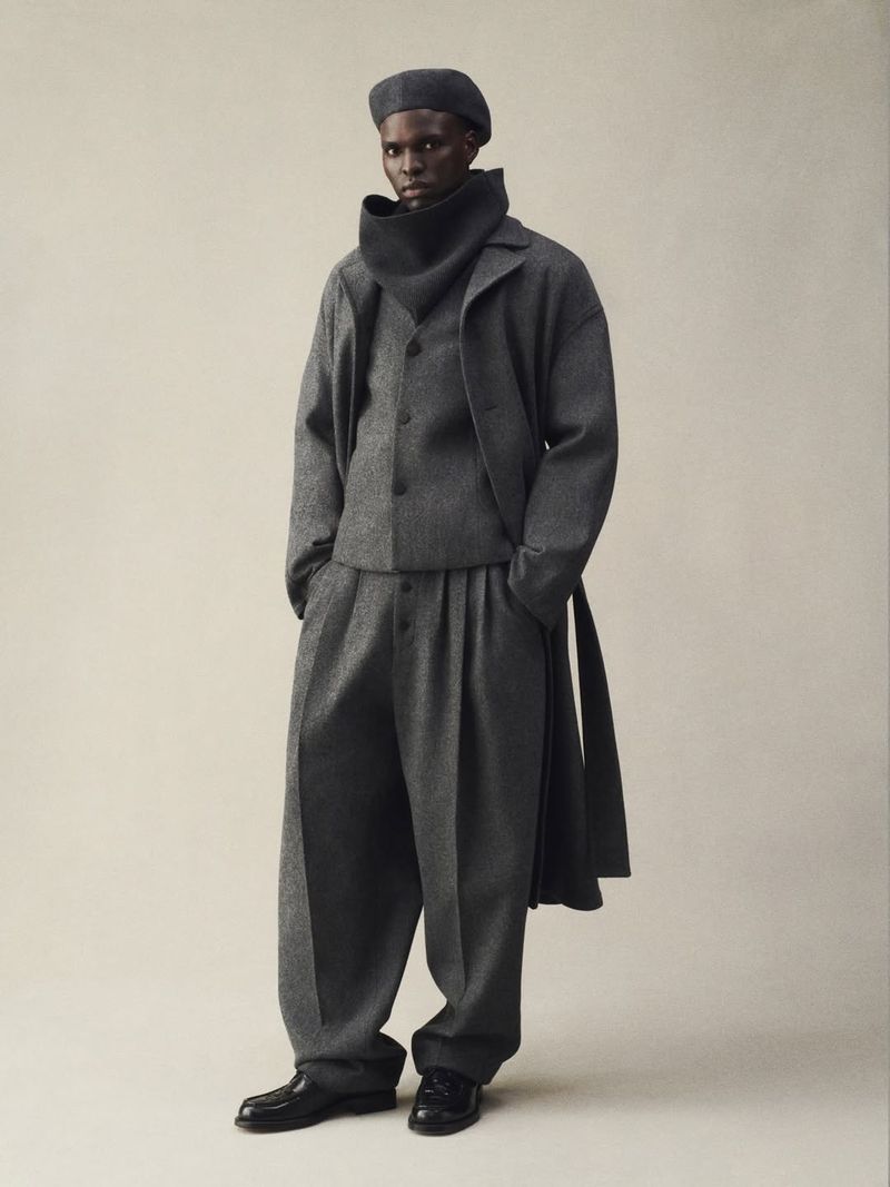 Studio shot of a dark-skinned male model in a grey wool suit with a beret. He poses in an indoor setting against a neutral background.
