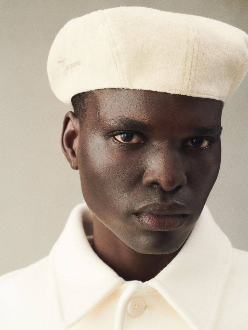 Close-up of a dark-skinned adult male fashion model in a white beret and matching shirt against a plain neutral background.