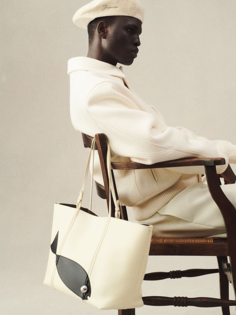 A man wearing a beret and a white outfit poses on a wood chair next to a light-colored tote bag with a black whale design.