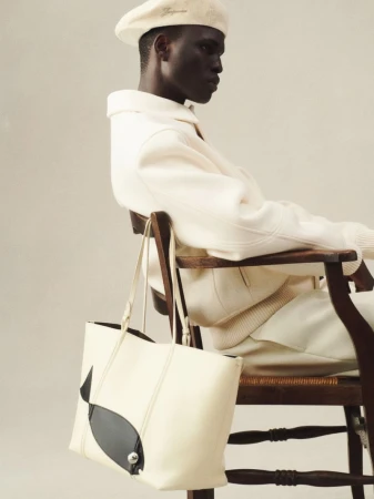 A man wearing a beret and a white outfit poses on a wood chair next to a light-colored tote bag with a black whale design.