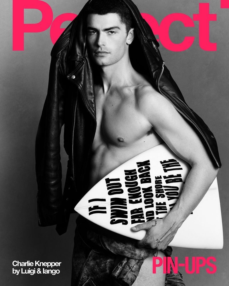Shirtless male model with surfboard and black leather jacket poses in a studio with pink text on a grey background for a magazine cover.