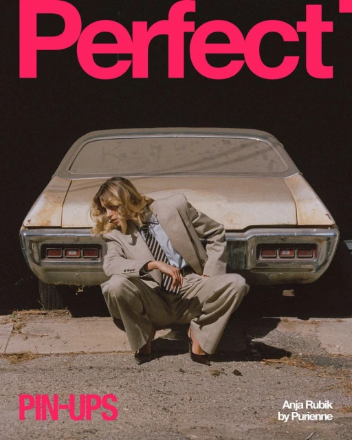 Fashion editorial cover shot: Blond woman in tan suit crouches in front of weathered classic car; black backdrop with pink magazine masthead.