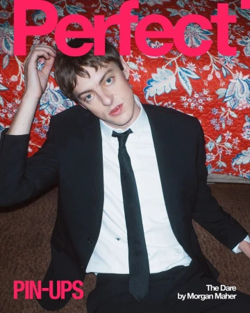 A young man in a black suit and tie sitting against a red floral patterned background for The Perfect Magazine.