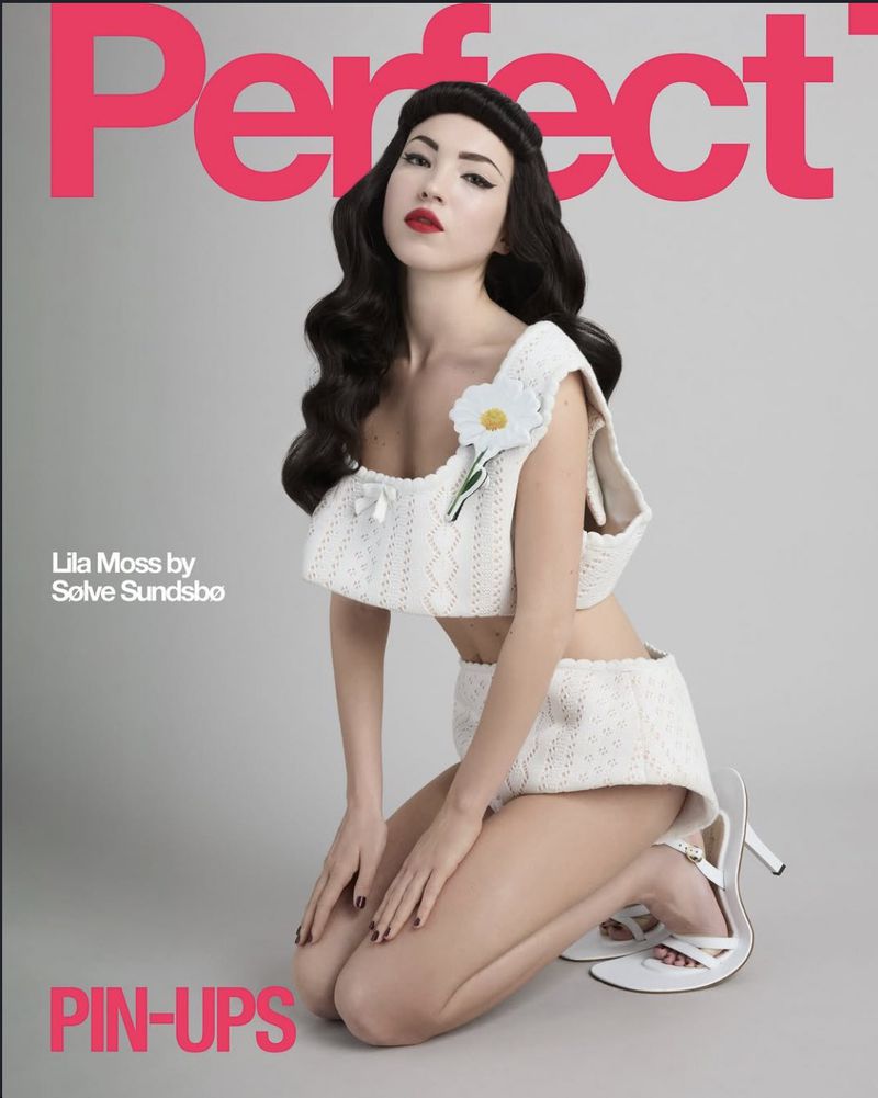 Lila Moss on a magazine cover, kneeling while wearing a white crocheted top and shorts. She's wearing white heels and has red lips.