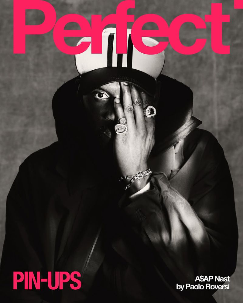 A black and white studio portrait of a dark-skinned man with hand jewelry covering his face, wearing a hat and jacket, with 'Perfect' text.