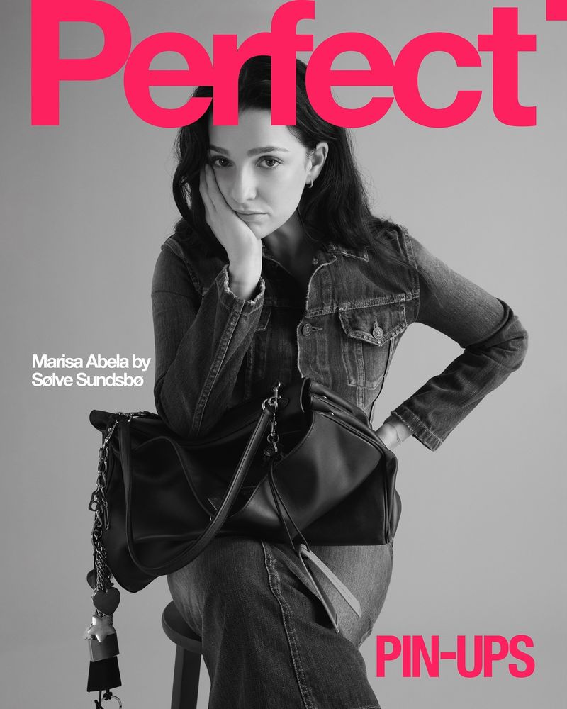Marisa Abela poses in denim jacket and jeans with a black handbag on a magazine cover with bright pink text and a grey background.