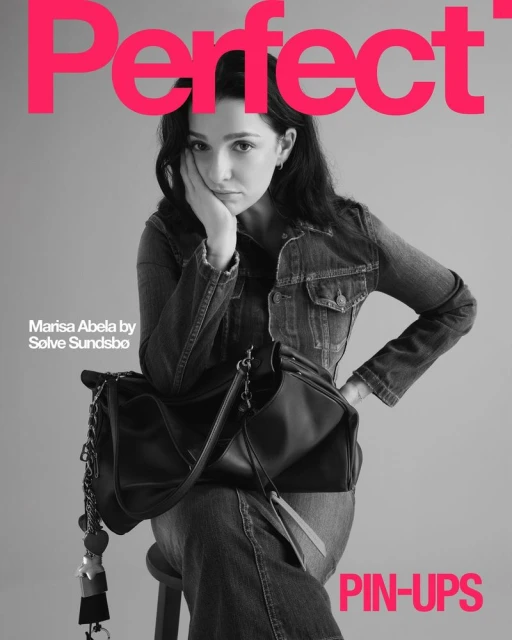 Marisa Abela poses in denim jacket and jeans with a black handbag on a magazine cover with bright pink text and a grey background.
