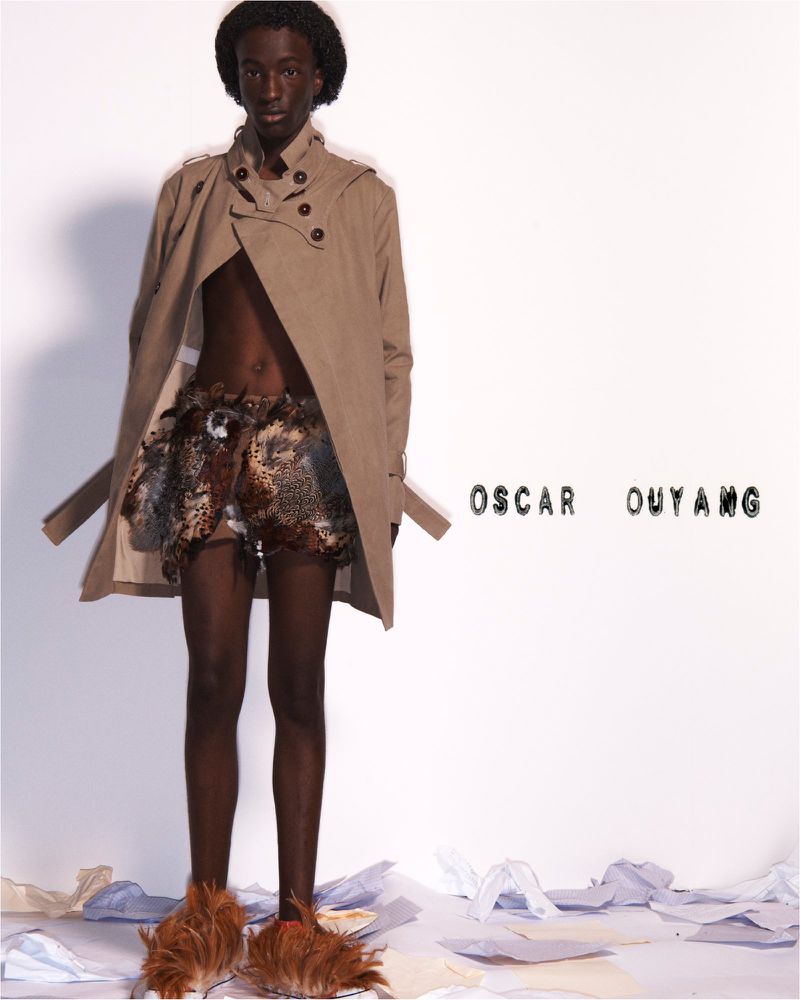 Fashionable Black male model showcases a beige trench coat and tribal-inspired shorts, paired with furry shoes, posing against a white background.